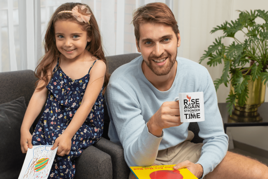 A smiling man holds a mug that reads “Rise Again Stronger Every Time” while sitting on a couch next to a young girl in a blue floral dress, who is holding her colorful drawing—perfect inspiration for motivational t-shirts for women. A plant sits nearby.