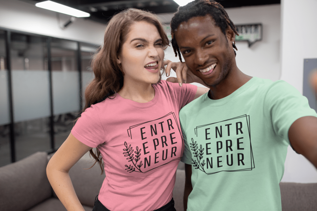 Two people smiling and posing together indoors, both wearing t-shirts that say ENTREPRENEUR in bold letters. One person is winking; both appear energetic and happy in a modern office setting.