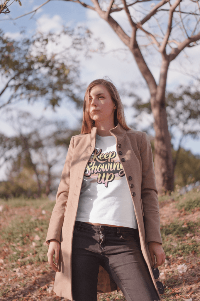 How Motivational T-Shirts Inspire Confidence Daily (+ Outfit Ideas) | Empowering T-Shirts by Quotnair Determined woman standing outdoors in nature, wearing a white motivational t-shirt that says "Keep Showing Up," layered under a tan coat, symbolizing persistence and inner strength.